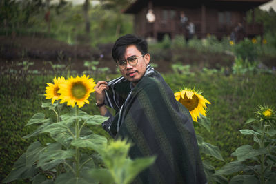 Portrait of young man standing amidst sunflowers blooming on field