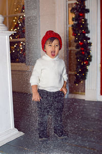 Boy stands on the porch of the house in a sweater and a red hat