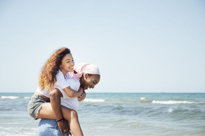 Delighted black mother piggybacking young daughter while having fun with closed eyes at seaside in summer