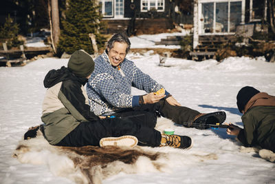 Smiling father talking with son while boy fishing lying on snow during sunny day