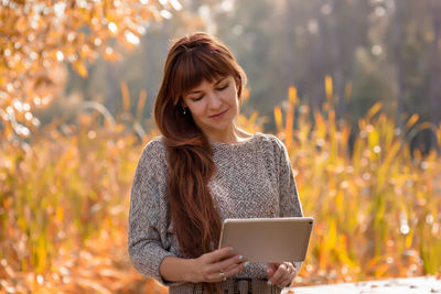 Portrait of  beautiful woman with long  hair, sitting in an autumn park, holding a digital tablet 