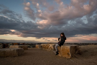 Young girl sits on a rock at the top of a hill at sunset