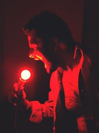 Side view of man shouting while holding illuminated light bulb in darkroom