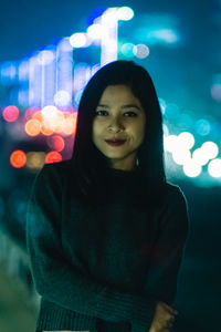 Close-up portrait of smiling young woman standing against illuminated light