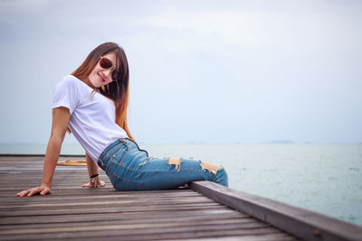 Young woman wearing sunglasses sitting on pier by sea against sky