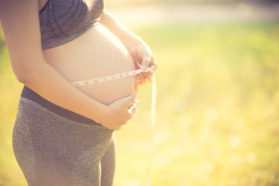 Midsection of pregnant woman measuring belly while standing on field