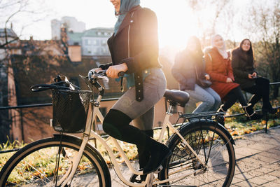 Low section of woman cycling by female friends sitting on railing in city