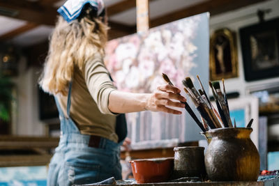 Low angle side view of female artist taking paintbrush from pot with various brushes while painting picture on canvas in art workshop