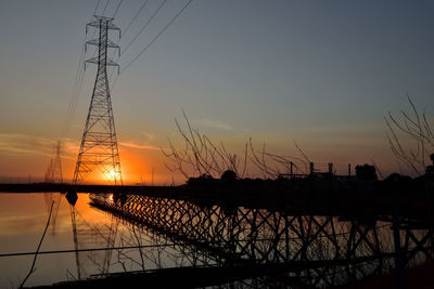 Silhouette electricity pylon by lake against sky during sunset