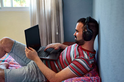 Young man works with his laptop in his bedroom, he is listening music