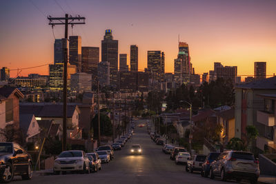 Vehicles on road amidst buildings in city against sky during sunset