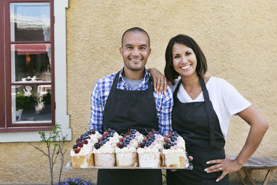 Smiling owners standing with tray of pastries outside cafe