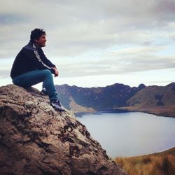 Man sitting on rock over lake against cloudy sky