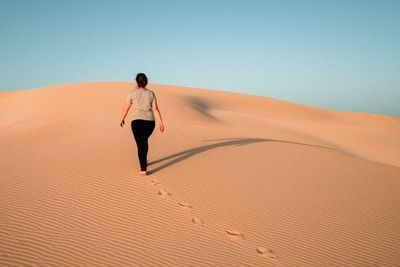 Rear view of woman walking on sand dune at desert against clear sky