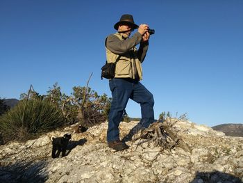 Full length of senior man photographing through digital camera on mountain against clear blue sky