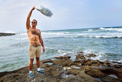 Full length of shirtless man holding his t-shirt while standing at beach against sky