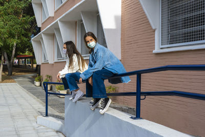 Female friends looking away wearing protective face mask while sitting on railing during sunny day