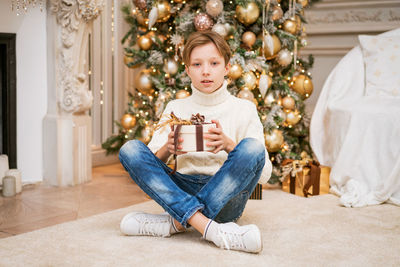 Cute boy in sweater sits by the christmas tree with gifts.