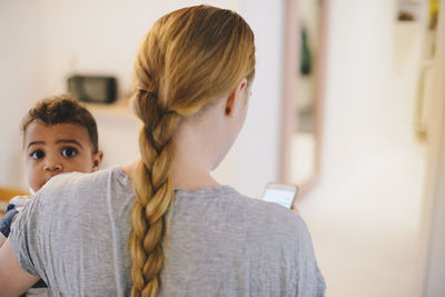 Rear view of mother with braided hair carrying son at home