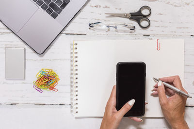 High angle view of woman writing on paper while holding mobile phone by laptop over table