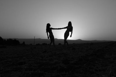 Silhouette women holding hands on landscape against sky during sunset