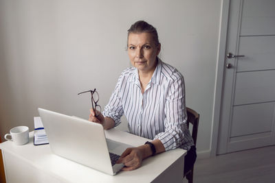 Woman in a white striped shirt aged sits at a computer at home with glasses