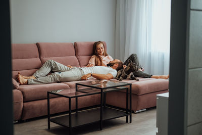 Cheerful woman and man relaxing on pink sofa with pet, couple having leisure with french bulldog