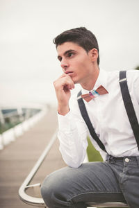 Young man with hand on chin looking away while sitting on railing against sky
