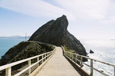 Footbridge leading towards mountain over sea against sky during sunny day