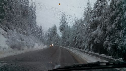 Road amidst trees seen through wet windshield in winter