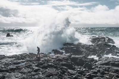 Full length of hiker against waves splashing on rocks at beach