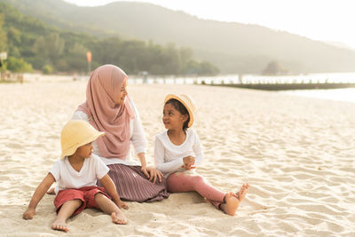 Mother wearing hijab with children sitting on sand at beach