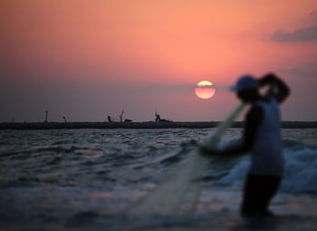 Man holding fishing net while standing in sea against sky during sunset