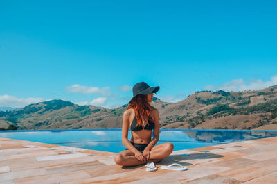 Woman sitting by swimming pool against blue sky