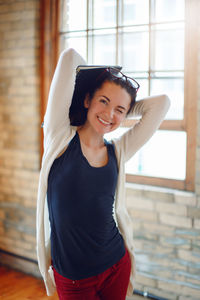 Portrait of smiling woman with hands behind head standing against window