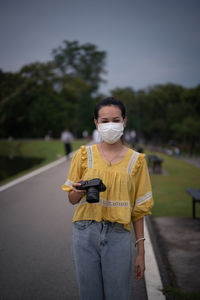 Young woman in yellow casual shirt hand holding camera and wear mask