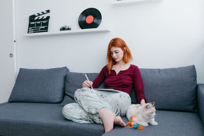 Young barefoot female with red hair browsing internet on tablet while sitting with cat on couch at home