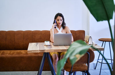 Focused female entrepreneur discussing business project on mobile phone while using netbook and working remotely at table in cafe