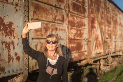 Adult woman wearing sunglasses taking a selfie with her smartphone of abandoned train cars.