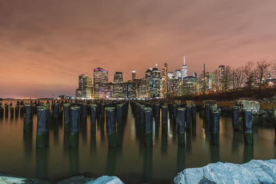 Illuminated wooden posts in sea against buildings during sunset