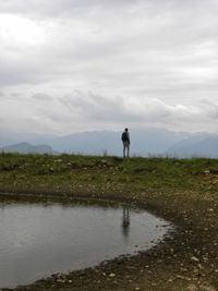 Rear view of man standing by pond at cliff against cloudy sky
