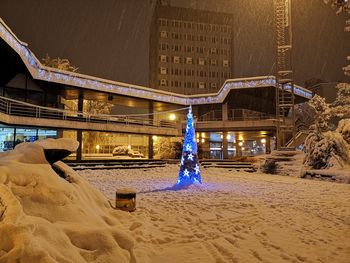 Illuminated street light on snow covered land by building at night
