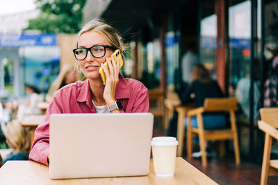Young working woman talking on the phone and using a laptop while sitting in a coffee shop