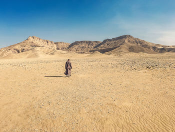 Rear view of man walking by mountains on desert against sky