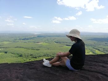 Side view of young woman sitting on landscape against sky