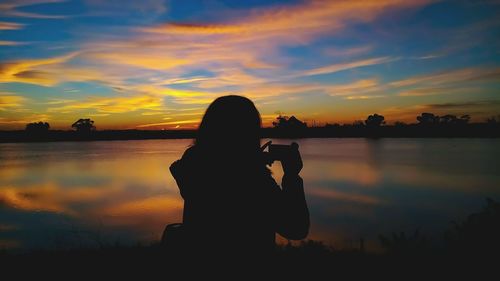 Silhouette woman photographing lake against sky during sunset