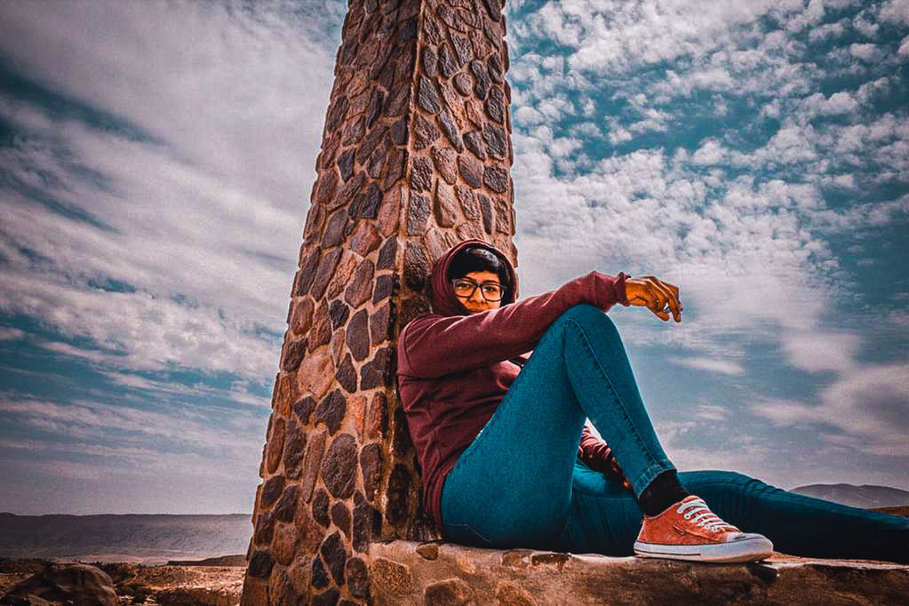 PORTRAIT OF YOUNG WOMAN SITTING ON ROCK AGAINST SKY