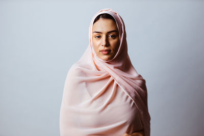 Portrait of young woman wearing hijab standing against gray background