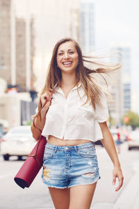 Portrait of smiling woman with exercise mat walking on road in city