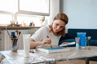 Woman artist painting picture at home with acrylic paints and spatula over the window. adult hobby